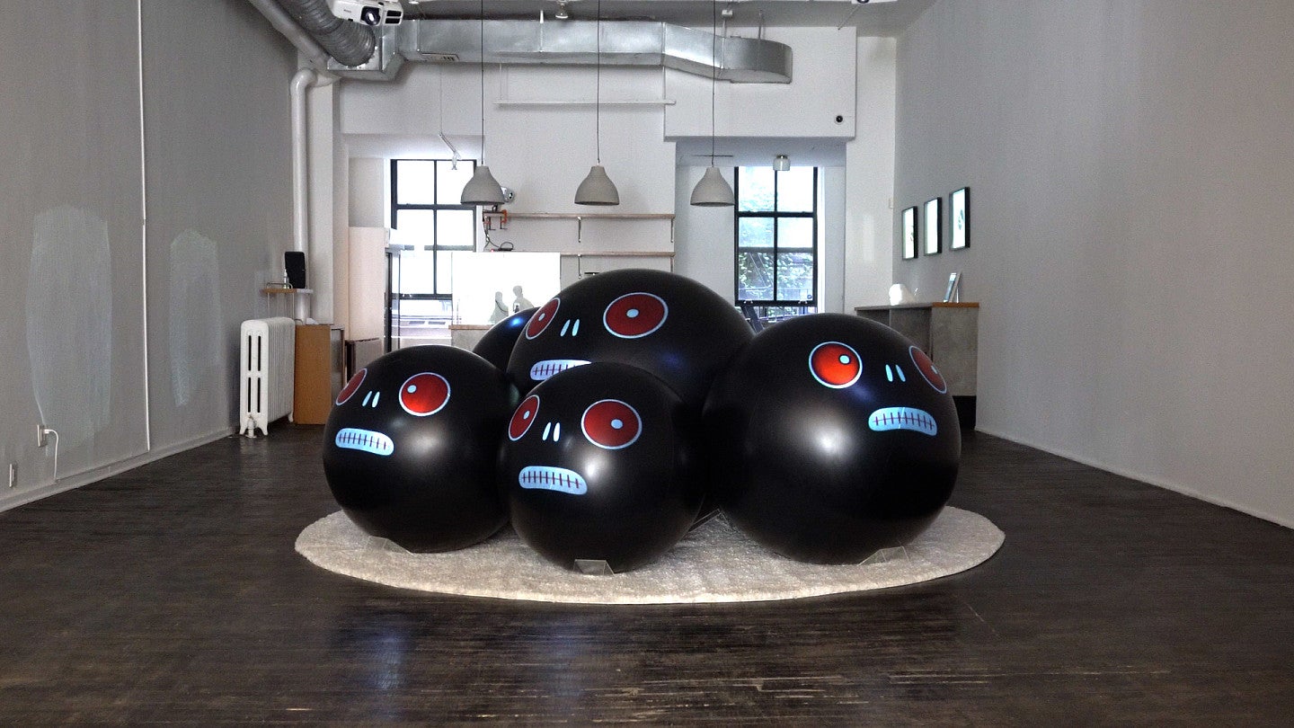 Sculpture of large black balls with cartoon-style faces. The faces have red eyes, 2 silver marks to make a nose, and a chrome oval with a stitch design to form the mouths.