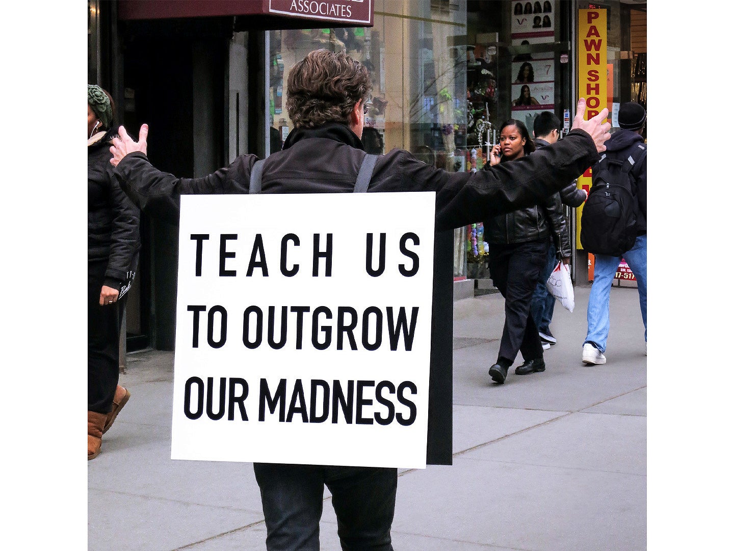 Image from Alfredo Jaar, titled "Teach Us to Outgrow Our Madness", 2018. Shows a man with a sandwich board on a city sidewalk with his back facing the camera. 