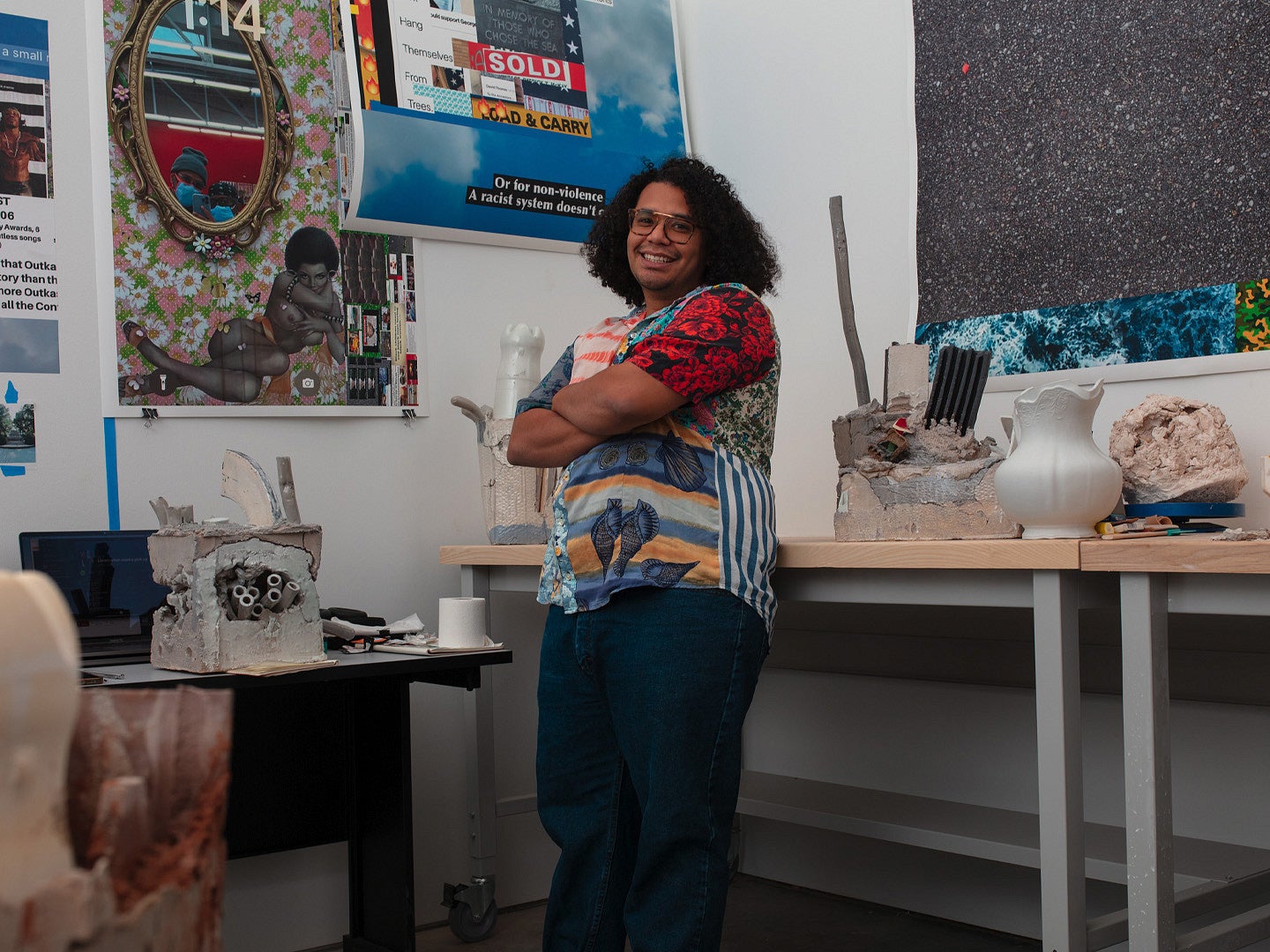 Photograph of artist, Kahlil Robert Irving. Photo shows a smiling person with neck length hair and a colorful shirt posing in an artist studio with pieces and other artworks on display. 