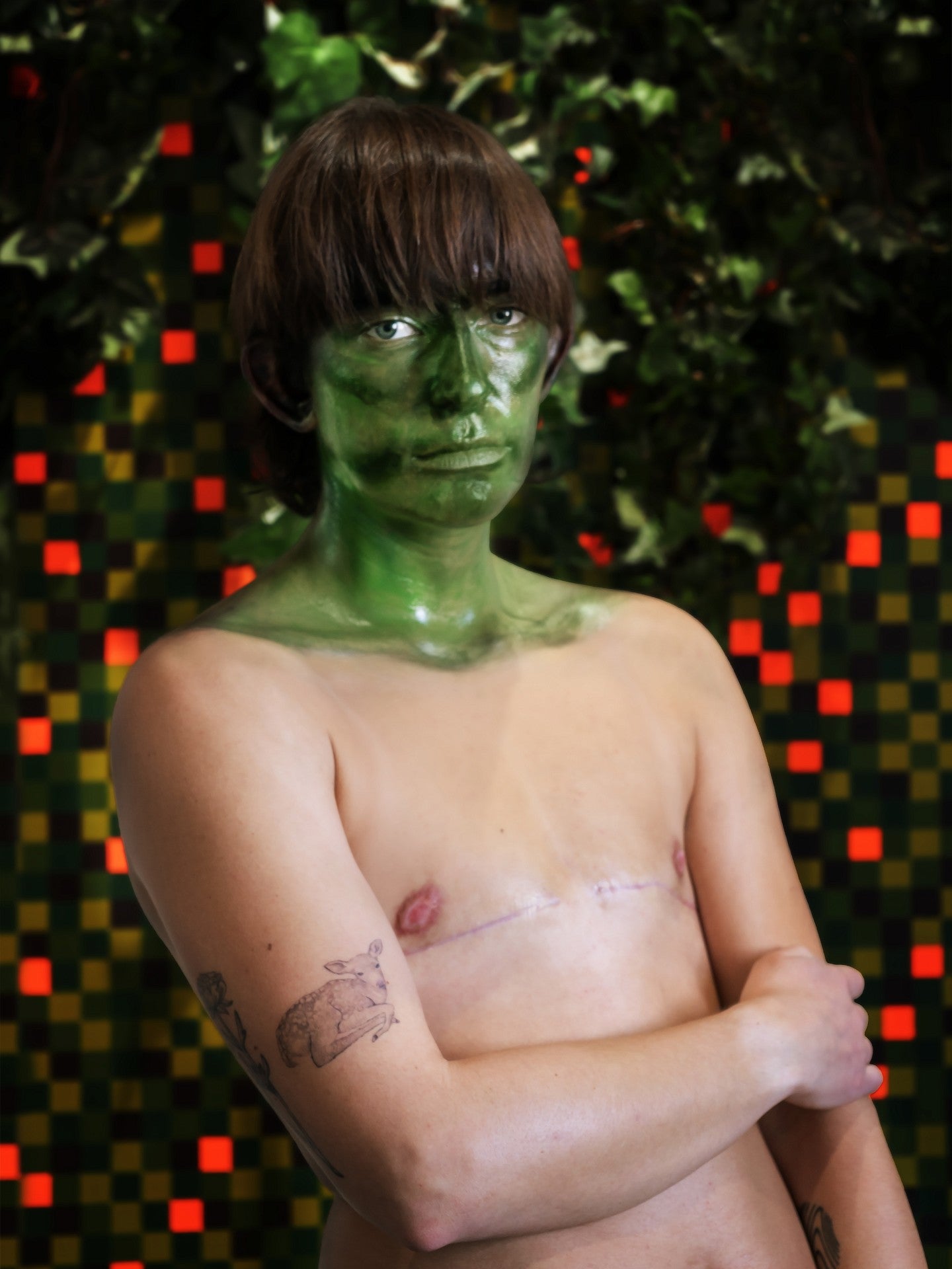 Photograph of the artist with green face paint in front of a multicolored abstract art piece.