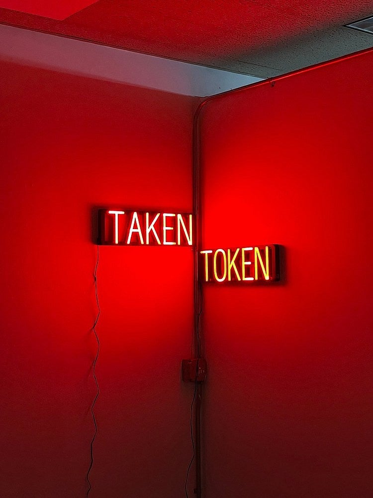 Photograph of two neon signs that say "Taken" and "Token". The ominous glow from the signs has lit up the white room and turned its walls red. 