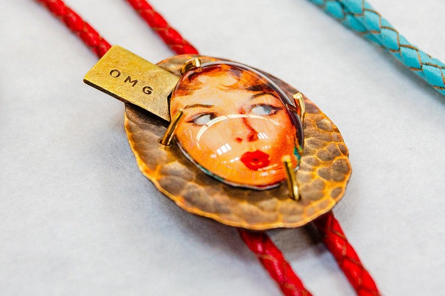 Jewelry Sale news header, shows a close up of a necklace with a vintage women's face surrounded by a round, metal frame that is reminiscent of reptile skin and corded with a red cord. 