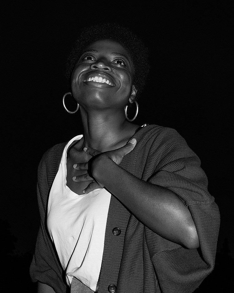 Black and white photo of a Black woman smiling up towards a point past the camera. Perspective is looking up. 