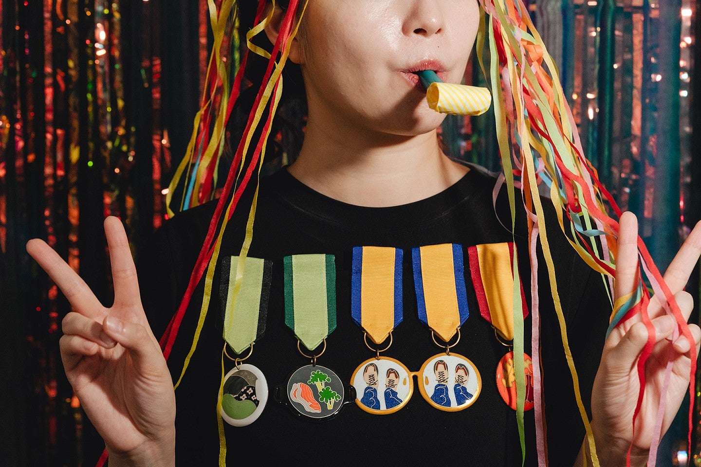 Photograph of an individual with a chest full of colorful medals, showing peace signs, with confetti on the head and a party favor in the individual's mouth. 