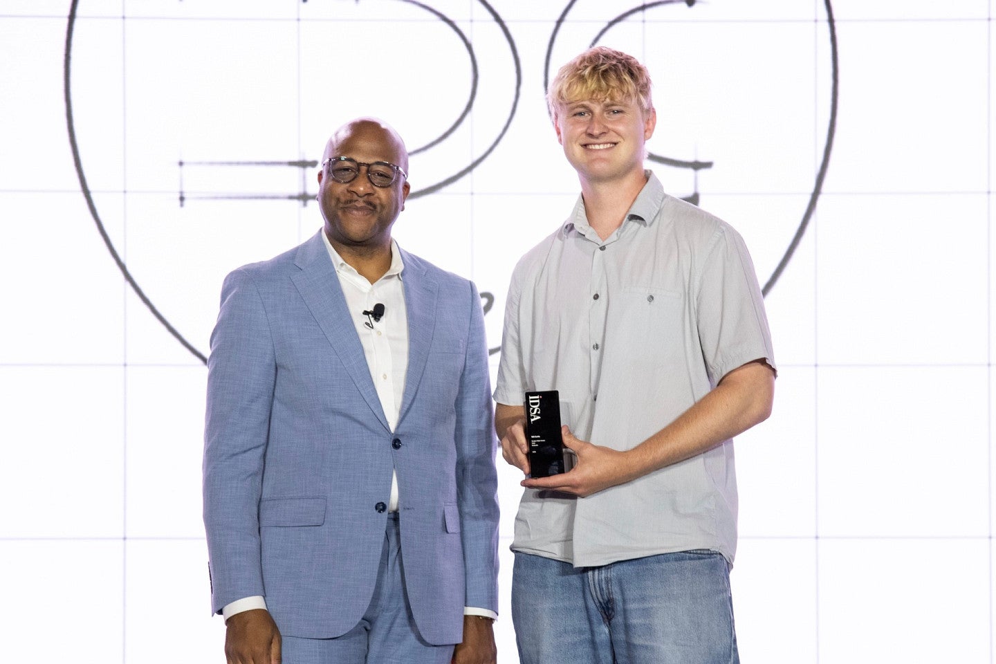 Will Curtis poses with IDSA member. Shows a young, tall, blond masc presenting person holding an award with a shorter, older bald man. 