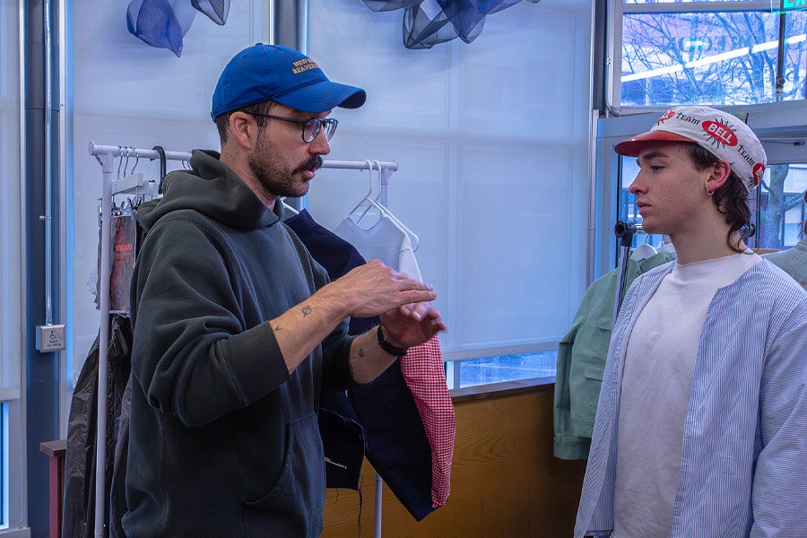 Tom Bonamici talks with a student about their design at a PD review. Shows two individuals in hats talking in front of clothing. 