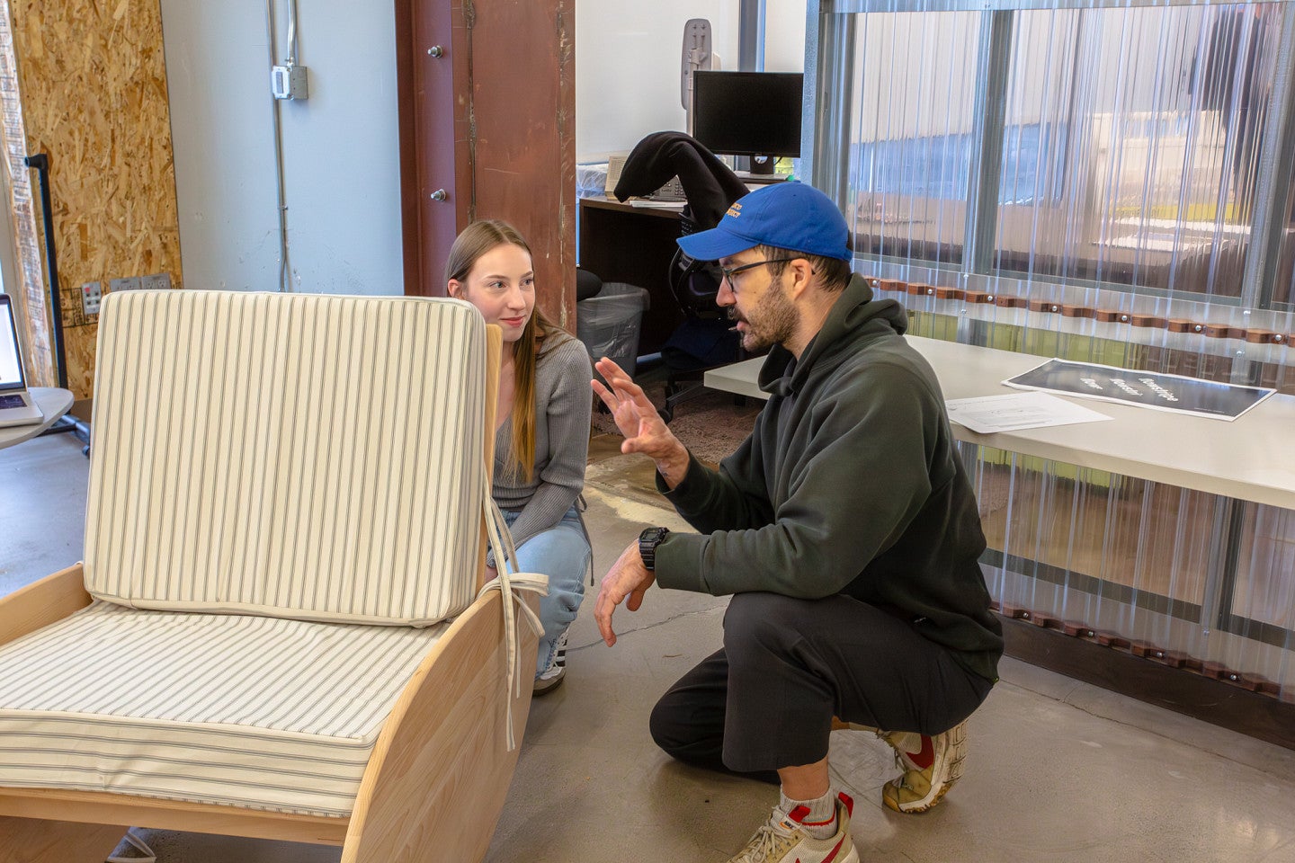 Tom Bonamici works with a student at a Product Design portfolio review. Shows two individuals behind a chair, squatting and talking. 