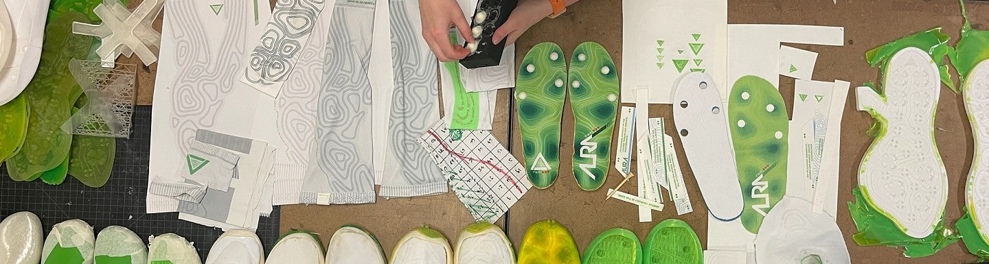 Photograph of green shoe inserts and shoe designs spread across a table with a student looking on. 