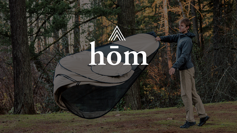 Man setting down a tan and black tent in the woods. He is wearing rain-resistant apparel.