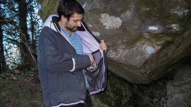 Man wearing a puffy, black jacket with a silky, pink hood and liner. The inner pockets are spacious enough to fit a larger water bottle. The man is in a wet, rocky environment.