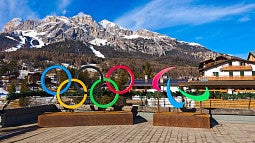News Header for Olympic Story. Shows the  Olympic rings and other designs in front of a scenic, snowy mountain range in Italy. 