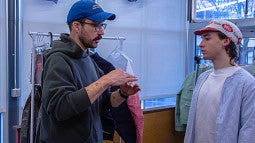 Tom Bonamici talks with a student about their design at a PD review. Shows two individuals in hats talking in front of clothing. 