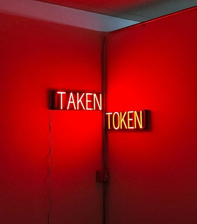 Photograph of two neon signs that say "Taken" and "Token". The ominous glow from the signs has lit up the white room and turned its walls red. 
