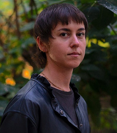 Headshot of the artist, Alice Bucknell. Shows an individual with short, cropped hair and neutral face in the woods looking into the camera. 