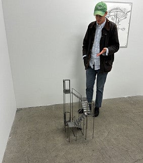 Photograph of the artist, Keith J. Varadi. Shows a masc presenting individual in a hat looking down at an art piece. 