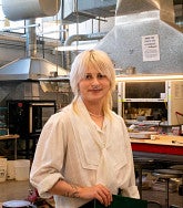 Photograph of a femme presenting person with long, blonde hair looking into the camera in a jewelry making classroom. 