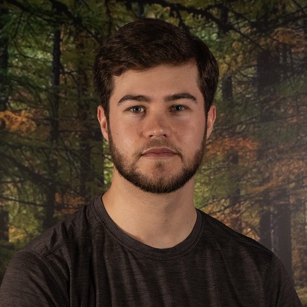 Masc presenting student stares into the camera in front of a wooded scene. 