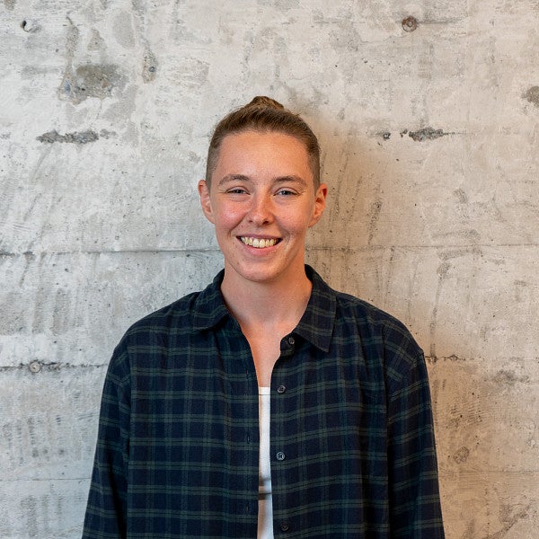 Shows an individual with their long hair pulled into a bun, smiling into the camera in front of a concrete wall.