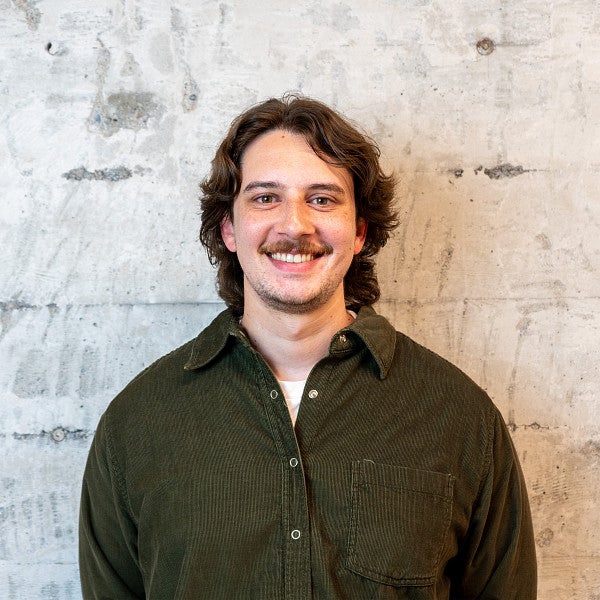 Masc presenting individual with neck length hair smiles into the camera in front of a concrete wall. 