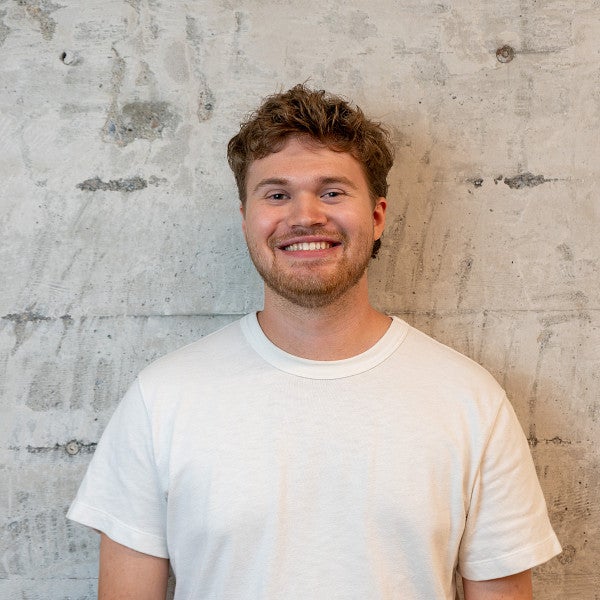 Masc presenting individual with short thick hair smiles in front of a concrete wall.