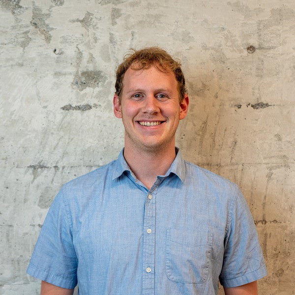 Masc-presenting individual with short hair smiles into the camera in front of a wall.