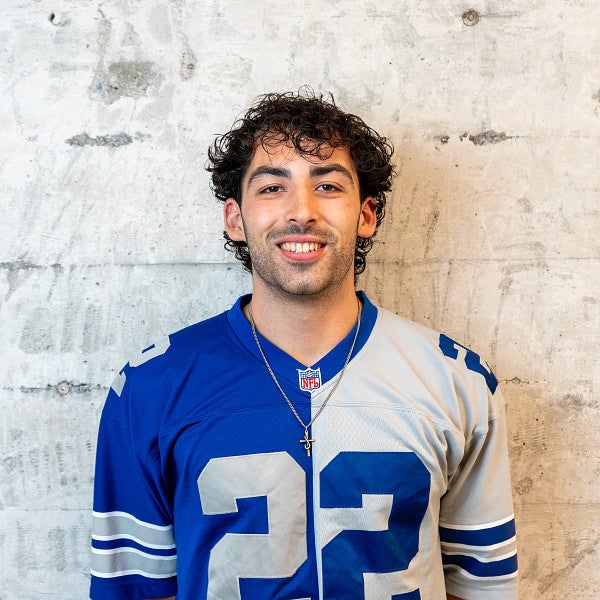 Masc presenting individual smiles into the camera in front of a concrete wall. 