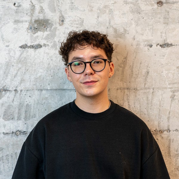 Masc presenting person with short, curly hair and glasses smiles into the camera in front of a concrete wall.