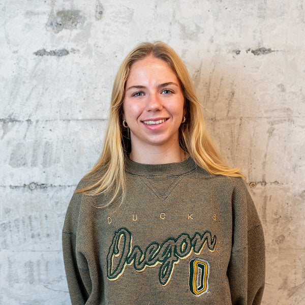 Femme presenting individual with shoulder length, blond hair smiles into the camera in front of a concrete wall. 