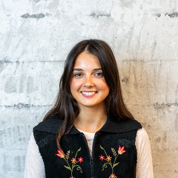 Femme presenting individual with long, dark hair smiles into the camera in front of a concrete wall. 