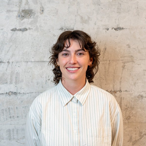 Femme presenting individual with shoulder length, wavy hair smiles into the camera in front of the wall. 