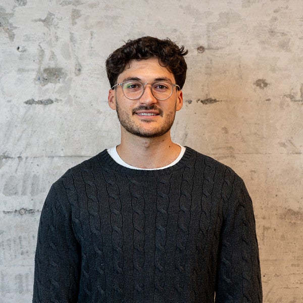 Masc presenting individual smiles into the camera in front of a concrete wall. 