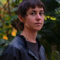 Headshot of the artist, Alice Bucknell. Shows an individual with short, cropped hair and neutral face in the woods looking into the camera. 
