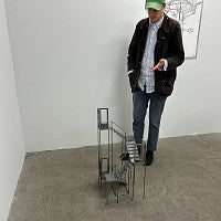 Photograph of the artist, Keith J. Varadi. Shows a masc presenting individual in a hat looking down at an art piece. 