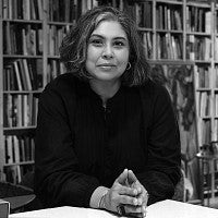 Photograph of artist Aruna D'Souza. Black and white photo of a femme presenting individual in front of shelves of books.